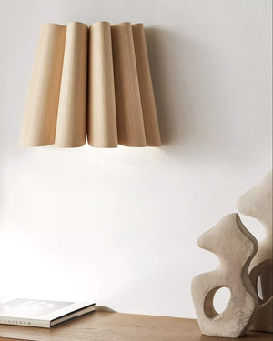 Wooden lamp with cylindrical design on a white wall, next to a book and abstract sculpture.