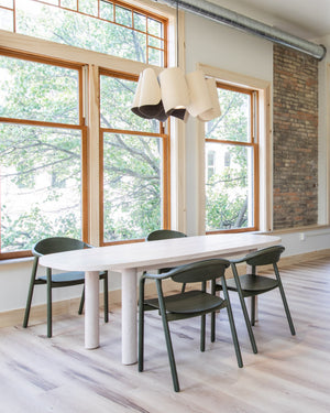 Modern dining area with wavy pendant on top of a long white table and green chairs in front of large windows.