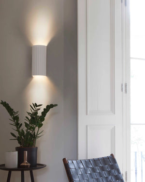 Modern wall light fixture on a neutral wall with a plant and chair in the foreground.