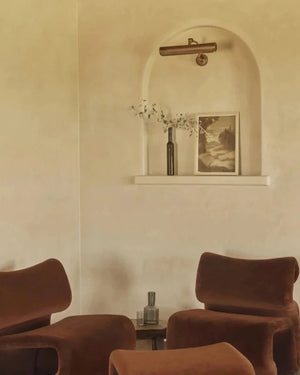 A solid brass picture light with an adjustable join, mounted on a wall above a picture, with two armchairs in the foreground.