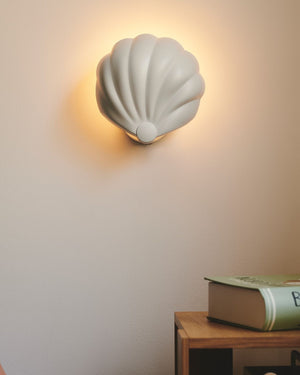 White shell-shaped wall light fixture on a plain wall with a book on a table in the foreground.