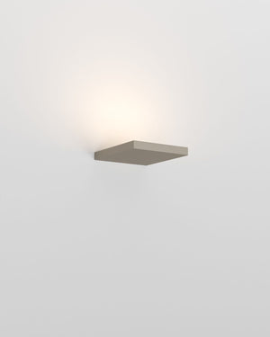 A minimalist rectangular wall sconce light fixture in a bronze finish, mounted on a plain white wall and casting a warm glow upwards.