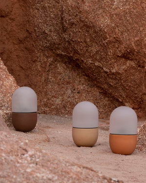 Three cylindrical lamps with different color combinations on a sandy surface with a rocky background