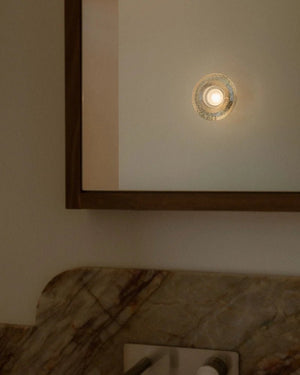 A warm, modern wall-mounted light fixture with a circular glass shade and glowing bulb, mounted on a beige wall above a marble sink with a brushed metal faucet.
