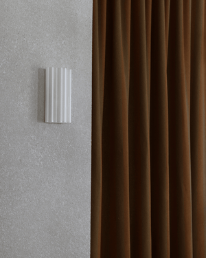 Fluted wall sconce mounted on a textured light gray plaster wall next to a floor-length rust brown curtain.