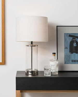 Lamp, bottle, and glass on a dark surface with a framed picture in the background.