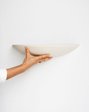 A hand holding a minimalist, semi-circular wall sconce in a warm off-white color against a plain white wall.