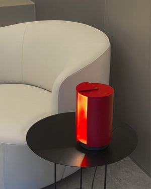Mid-century Red cylindrical lamp on a black side table next to a white curved chair.