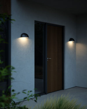 Two black dome-shaped outdoor wall sconces mounted on a light gray stucco wall on either side of a dark wood entry door at dusk.