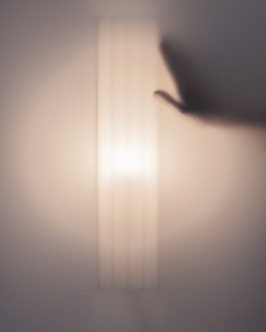A soft, abstract photograph of a glowing rectangular light fixture on a warm wall, with a hand silhouette 