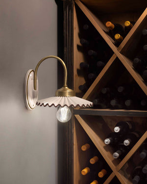A wall-mounted sconce with a curved brass arm, a white scalloped shade, and a clear bulb, shown lit next to a wooden wine rack filled with bottles.