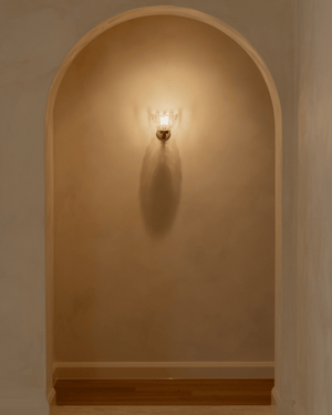 A warm floral wall sconce with a gold-toned brass fixture and clear glass shade, mounted on a beige plaster wall inside an arch.