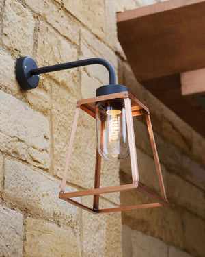 Wall-mounted outdoor light fixture with copper frame and black arm on a stone wall.