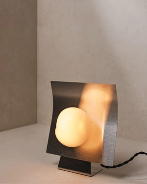 Glass and stainless steel table lamp with curved brushed panel and glowing handblown glass globe in neutral interior corner