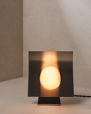 Front view of a glass and stainless steel table lamp with curved brushed panel and glowing handblown glass globe in neutral interior corner
