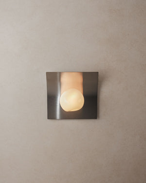 Glass and stainless steel wall light with curved brushed plate and glowing handblown glass globe on neutral plaster wall