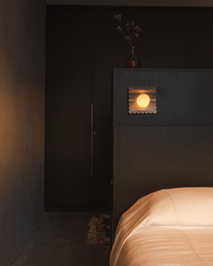 Wave Wall Light glowing above a dark timber bedhead beside a bed with peach linen