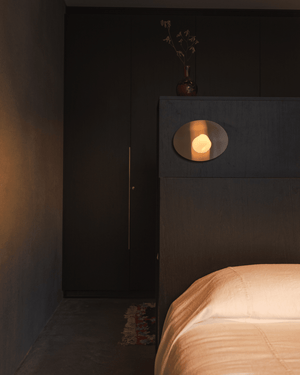 Wink Wall Light glowing above a dark timber bedhead beside a bed with peach linen