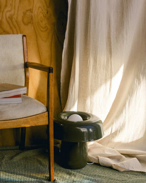 Wooden chair with a white cushion next to a greenlamp on a textured surface with a curtain in the background.