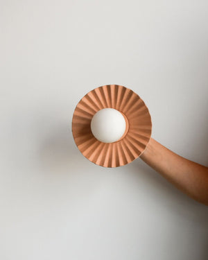 Hand holding a pleated leather shade wall light with a glass orb in the middle in front of a white background