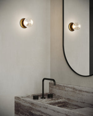 Handmade Sora Clear Glass Wall Sconce in a bubble glass finish with warm light on above a modern bathroom sink below a curved mirror