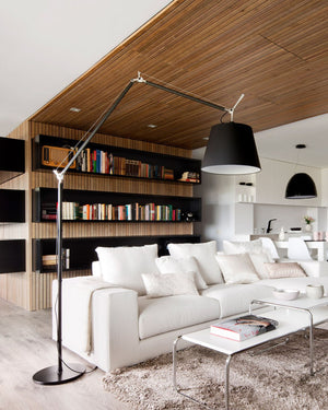 A modern living room featuring a white sectional sofa, a large black floor lamp with an adjustable arm, a white coffee table, and a slatted wood wall with built-in shelves and books.