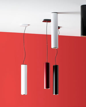 Three modern cylindrical pendant lights hang from a white ceiling against a bold red wall. The shades are in white, dark red, and black, suspended by thin black cords.