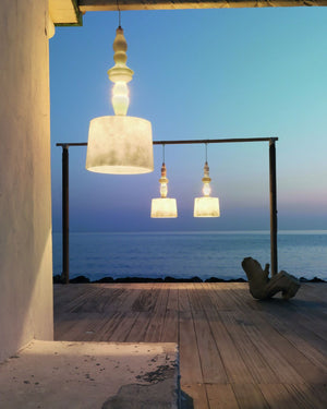 Three big Italian Design Outdoor Pendant lights with textured off-white drum shades and warm glowing bulbs hang over a wooden deck at dusk, overlooking a calm ocean and sky.