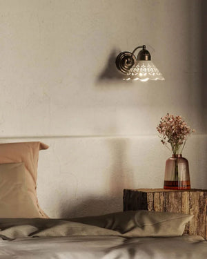 A wall-mounted sconce with a textured glass shade and dark metal fixture, glowing above a bed in a softly lit bedroom. 