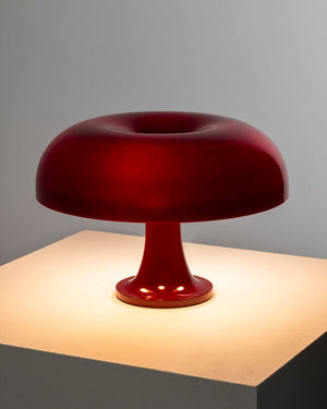 A glossy deep red table lamp with a mushroom-shaped shade and pedestal base, illuminated from within and placed on a lit square pedestal against a gray background.