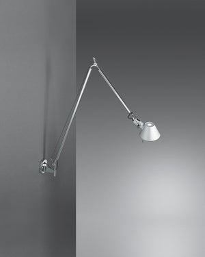 A modern wall-mounted adjustable arm lamp with a silver metal articulated arm and conical shade, shown against a dark gray wall with light casting on the right.