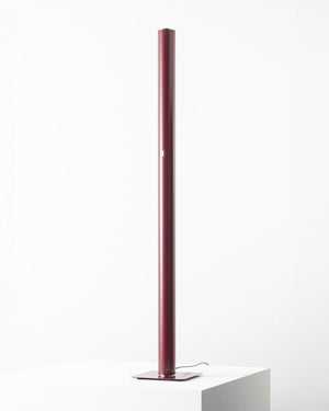 A modern minimalist floor lamp with a tall, slim cylindrical column in a dark red color, standing on a square metal base atop a white plinth against a clean white background.