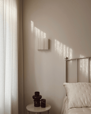 A modern wall-mounted light fixture with vertical pleated detailing, shown in a softly lit bedroom setting next to a bed and side table.