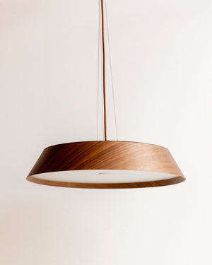 A modern pendant light with a wide, shallow conical shade made of dark brown wood on the outside and a white polycarbonate diffuser on the inside, suspended by a thin cord against a plain off-white wall.