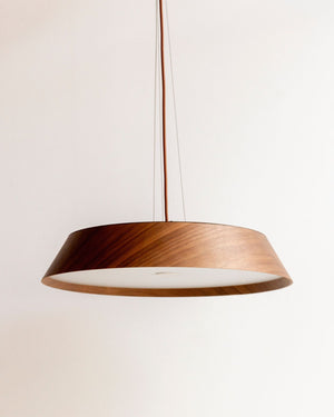 A modern pendant light with a wide, shallow conical shade made of dark brown wood on the outside and a white polycarbonate diffuser on the inside, suspended by a thin cord against a plain off-white wall.