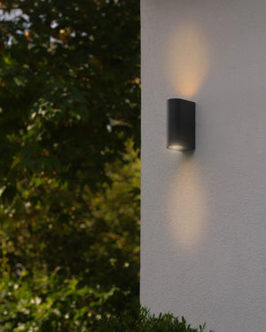 Black cylindrical outdoor wall light with dual up and down illumination on a white wall with greenery in the background