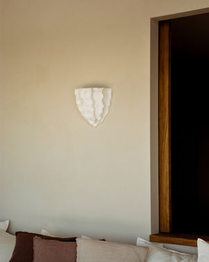 Decorative wall light fixture on a beige wall with pillows on a bed in the foreground.