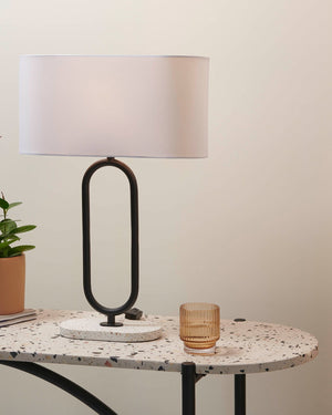 Oval Satin Black Table lamp with a white shade on a round marble table next to a plant and glass.
