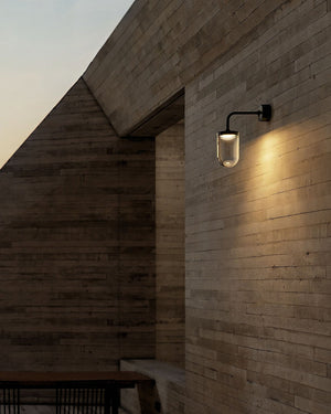 A modern black wall sconce with a clear cylindrical glass shade, mounted on a brick wall and casting a soft glow.