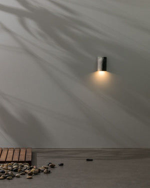 Wall-mounted outdoor light fixture with pebbles and wood at the base.