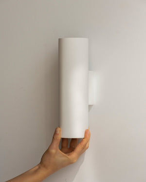 An up and down plaster pillar wall light tricolour being held by a hand against a plain white wall.
