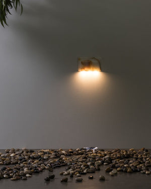 Wall-mounted light fixture casting a warm glow on a gray wall with pebbles at the base.