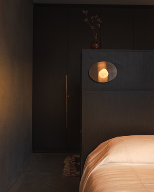 Wink Wall Light glowing above a dark timber bedhead beside a bed with peach linen